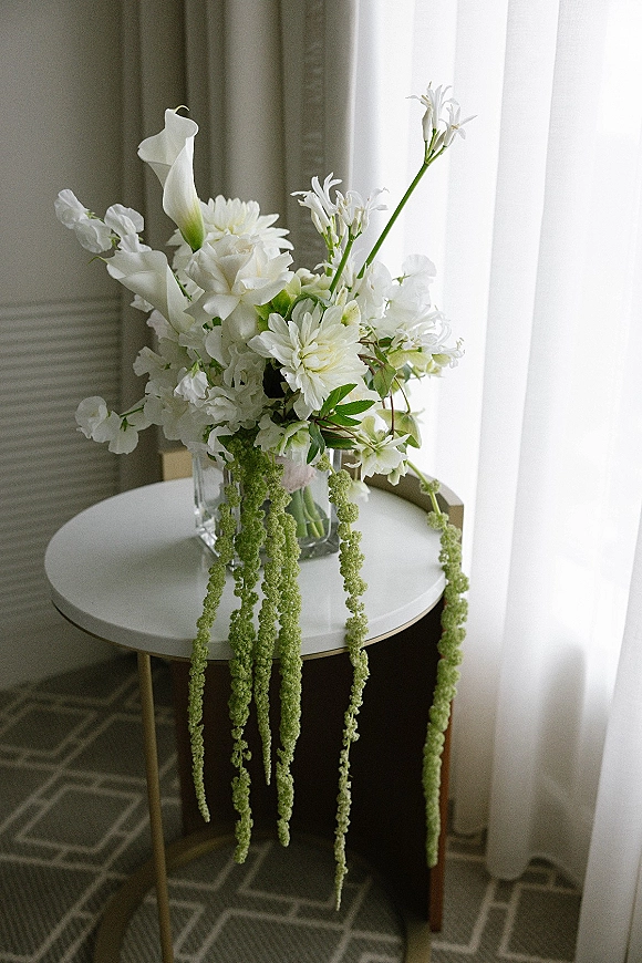 Wedding bouquet with white bridal bouquet blooms and cascading greenery in a glass vase on a side table by sheer-curtained window light