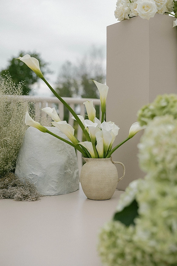 Wedding floral decor with calla lily arrangement in a ceramic pitcher on a pedestal plinth, draped fabric and moss on an outdoor terrace