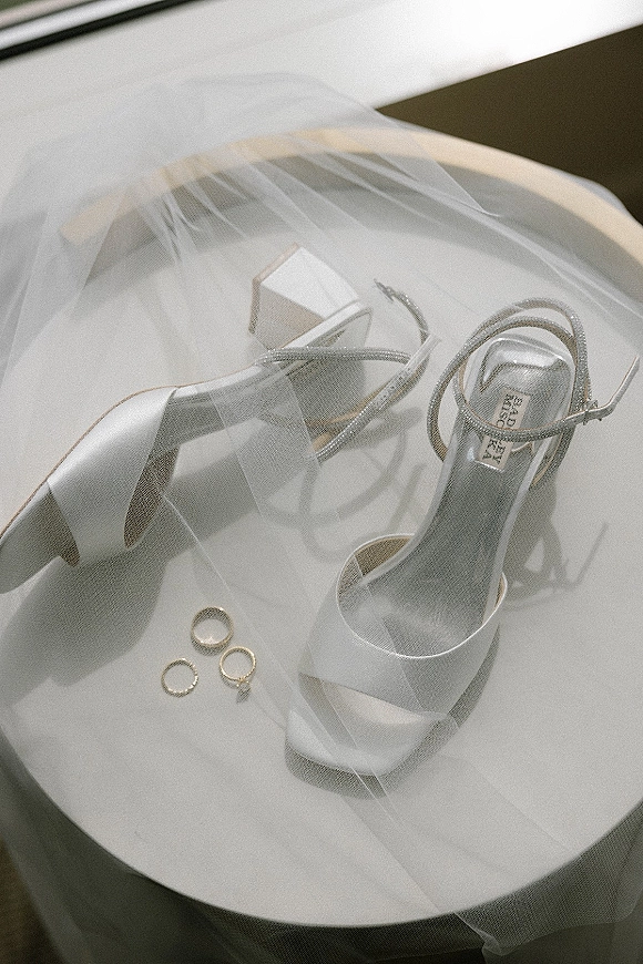 Bridal shoes in a white bridal heels flatlay with wedding rings and veil on a sunlit white tabletop, casting soft shadows