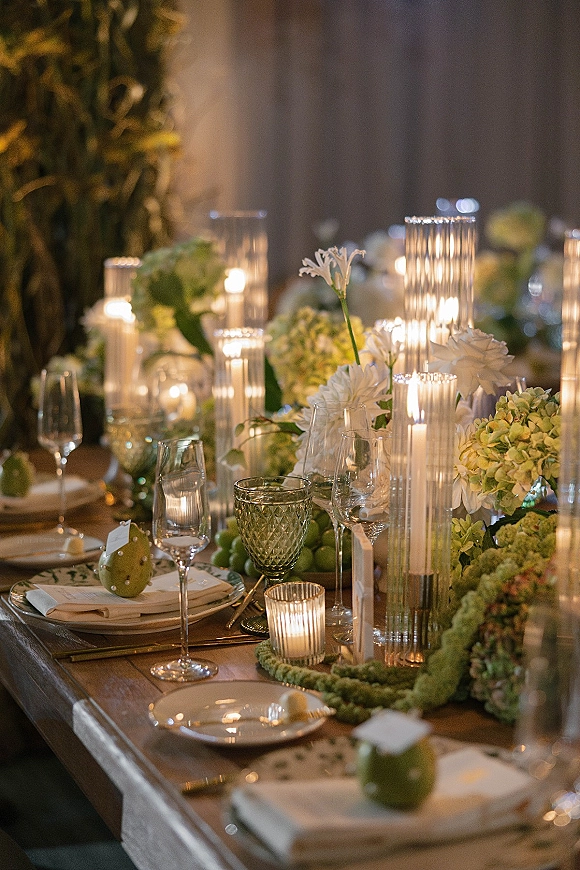 Reception tablescape with a candlelit wedding table featuring hydrangeas, white florals, green grapes, and ribbed glass holders on wood table backdrop