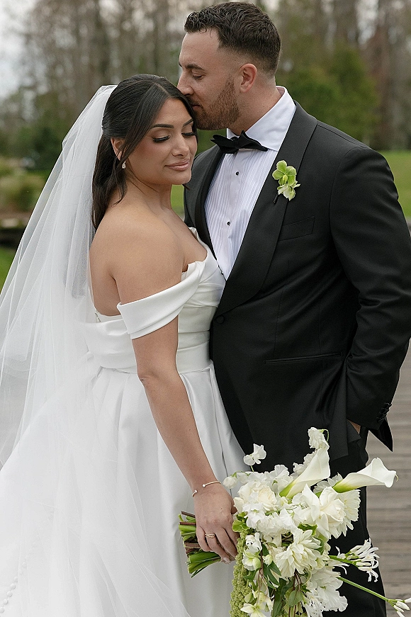 Couple portrait of bride and groom embrace with a forehead kiss, her veil flowing and white calla lily bouquet on a tree-lined lawn walkway
