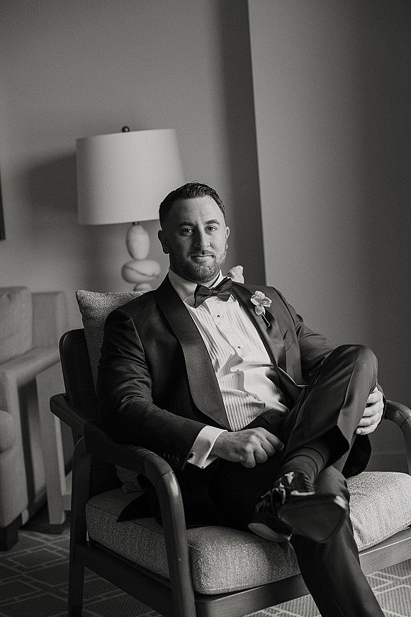 Groom portrait of a groom sitting in chair wearing a black tuxedo with bow tie and boutonniere in a hotel room, lit by a table lamp