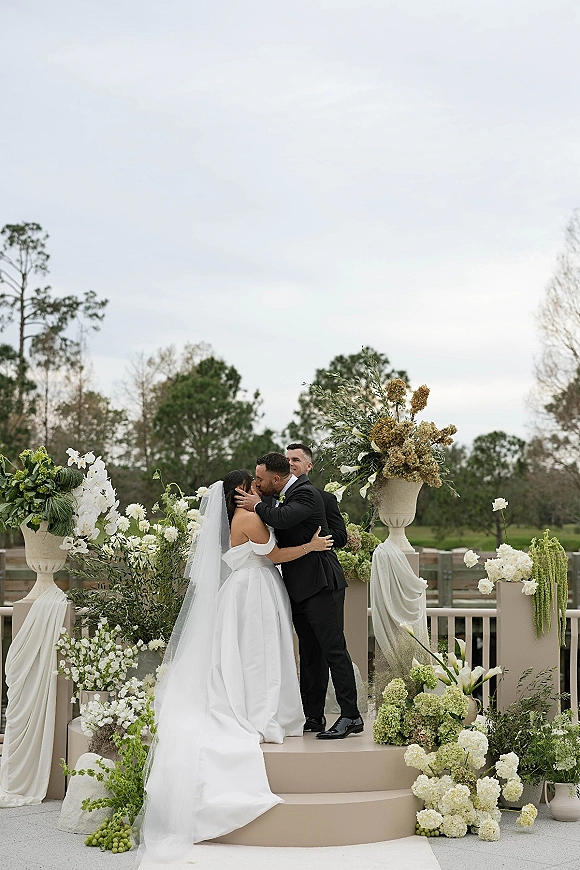 Ceremony kiss as bride in wedding dress and cathedral veil kisses groom in tuxedo beside white floral urns on outdoor terrace steps