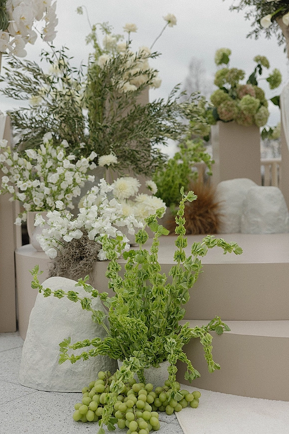 Ceremony floral decor with wedding floral pillars of white flowers and greenery on beige plinths, accented by green grapes on an outdoor terrace
