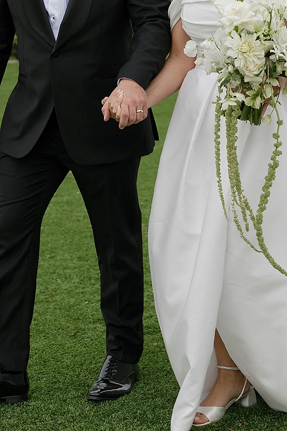 Wedding couple portrait of bride and groom holding hands, highlighting wedding rings, black tuxedo, satin gown, and white bouquet on lawn