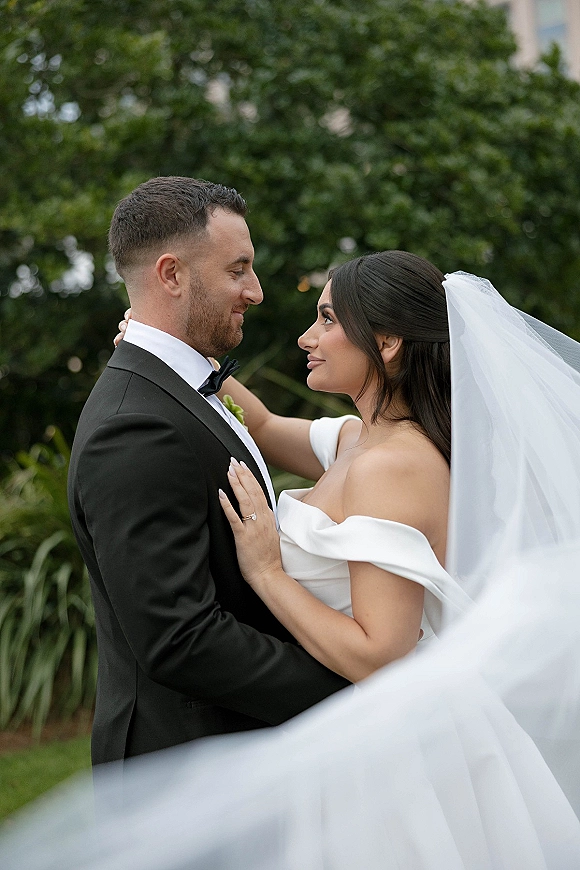 Couple portrait of bride and groom embrace in side profile, veil blowing across the foreground, with green garden trees behind them