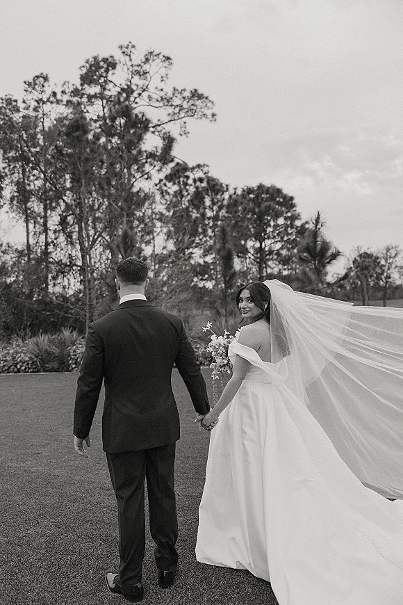 Couple portrait in a black and white wedding photo, bride and groom holding hands walking on a lawn as her long veil streams behind