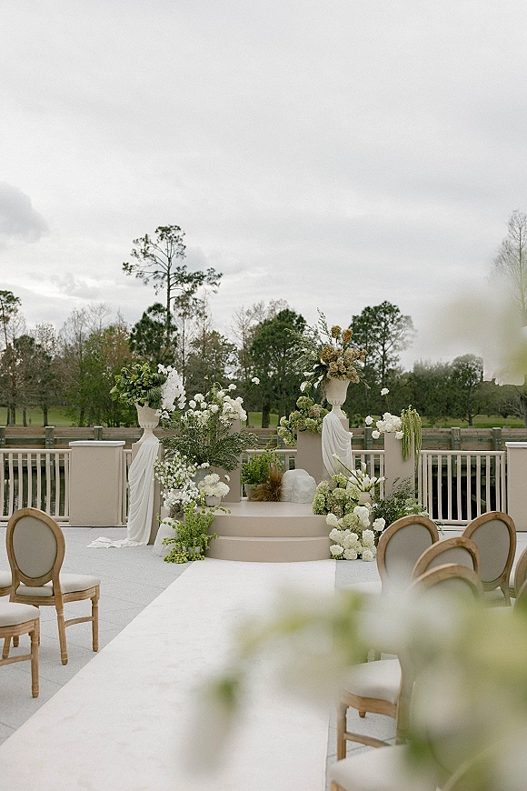 Ceremony setup with a white aisle runner leading to a tiered platform with pedestal urn florals and draped fabric on an outdoor deck