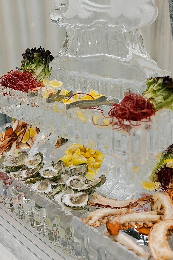 Seafood buffet display with wedding seafood bar oysters, crab legs and shrimp on ice with an ice sculpture, lemon wedges, and white drapery backdrop