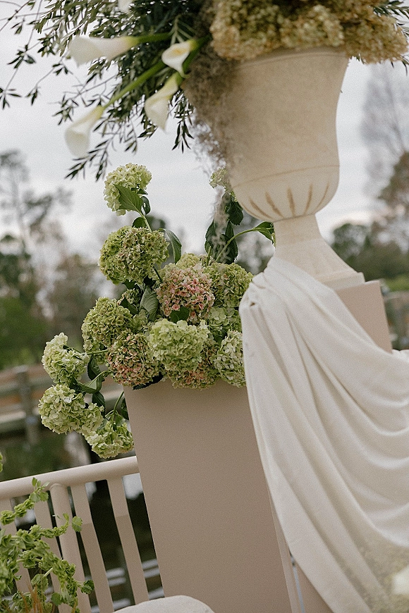 Ceremony floral decor with hydrangea ceremony flowers on urn and plinth pedestals, white draped fabric and hanging greenery on a balcony