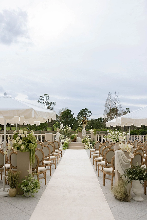 Ceremony setup for outdoor wedding ceremony with a white aisle runner, pedestal urn florals, and draped fabric on a terrace under umbrellas