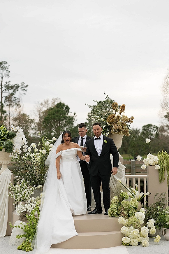 Wedding recessional as bride and groom walk the aisle holding hands, bride laughing in veil beside tux groom on terrace with florals