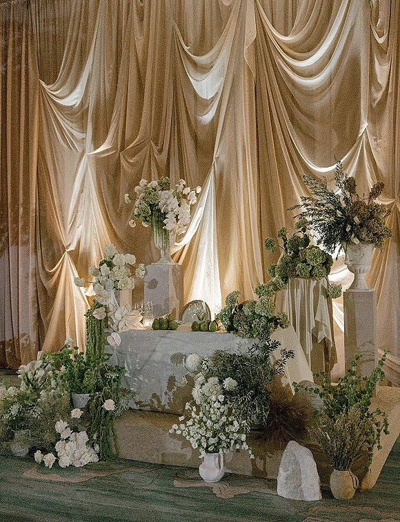 Reception tablescape with wedding sweetheart table styling, green and white florals, candles and fruit centerpiece against uplighted draped backdrop
