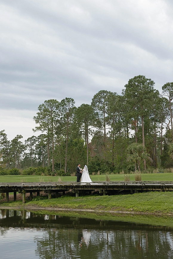 Wedding couple portrait of bride and groom on a wooden bridge by a pond, holding hands, her long veil trailing under cloudy sky