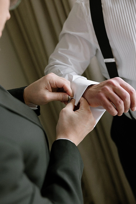 Groom getting ready, putting on cufflinks on a white dress shirt with suspenders, standing by softly lit curtains on wedding morning