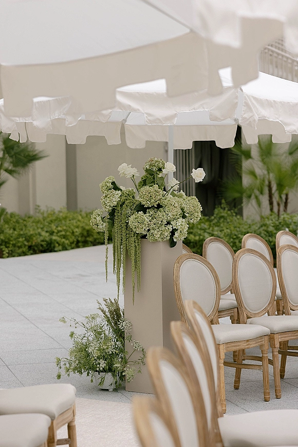 Ceremony aisle decor with white umbrellas shading green hydrangea and white rose florals on a stone patio lined with chairs and hedges