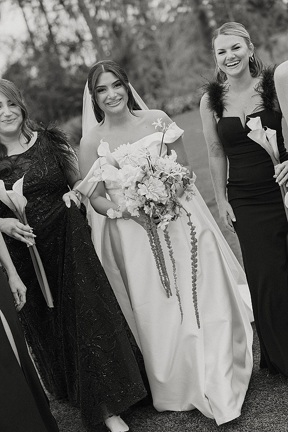 Bride with bridesmaids portrait on an outdoor lawn, bride in off-the-shoulder dress and veil holding a calla lily bouquet with trees behind
