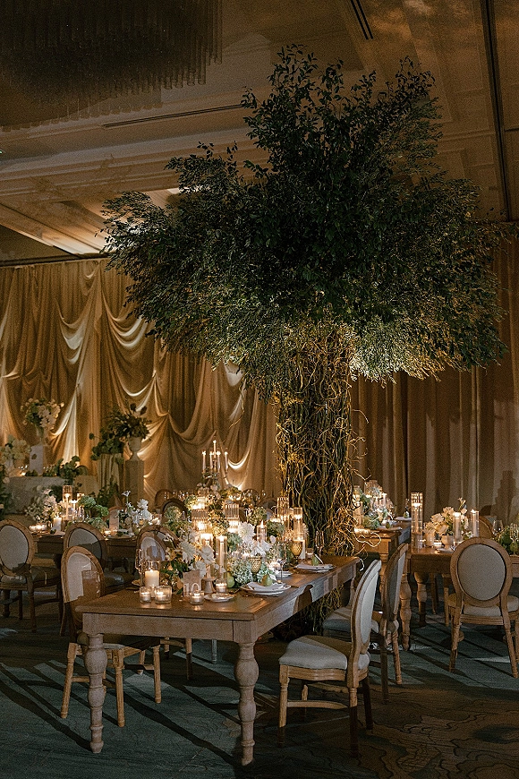 Reception tablescape with a wedding reception centerpiece tree over wood farm tables, taper candles and white florals beneath chandeliered draping