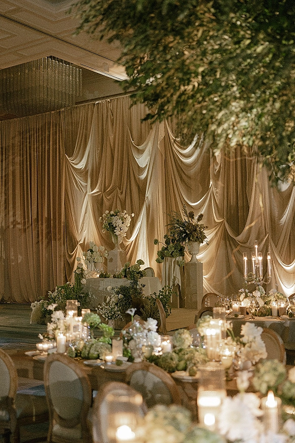 Reception stage decor with wedding drape backdrop, white florals and greenery on plinths, plus candlelit centerpieces beneath hanging greenery