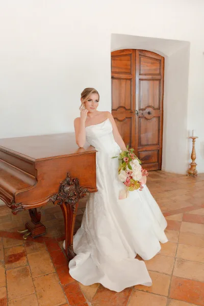 Bridal portrait beside a grand piano, the bride stands gracefully in a simple indoor setting, her posture calm and elegantly composed.