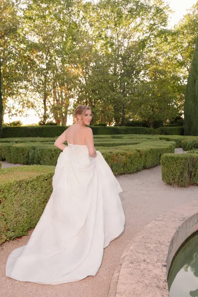 Bridal portrait of a bride with a long train, glancing over her shoulder on a gravel path by a stone fountain amid formal garden hedges.