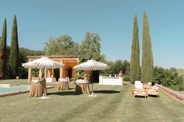 Cocktail hour setup shaded by scalloped umbrellas on a lawn beside a pool, framed by cypress trees and a stucco pavilion under blue sky.