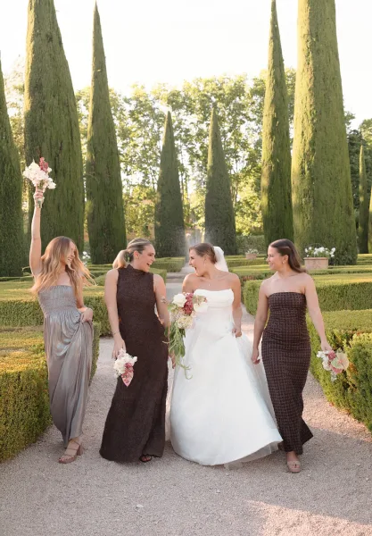 Bride and bridesmaids, a bridal party walking beneath cypress trees, carry bouquets along a gravel garden path lined with manicured hedges.