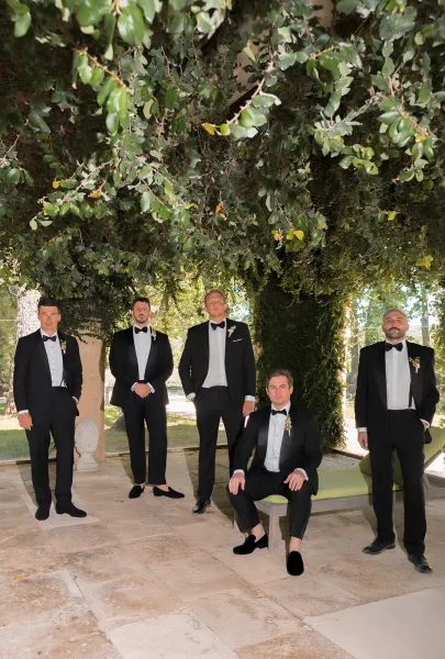 Groom and groomsmen in black tie wedding attire line up around a seated groom on a lounge chair under a leafy canopy on a stone patio.