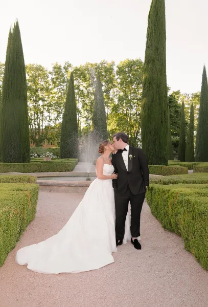 Wedding kiss portrait of a bride and groom in black-tie, his boutonniere framed by a fountain and cypress-lined formal garden.