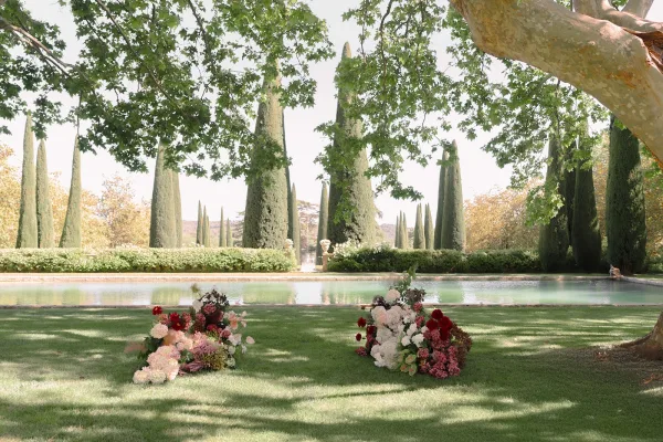 Ceremony setup featuring outdoor ceremony decor and ground floral arrangements by a reflecting pool, framed by cypress trees and garden hedges.