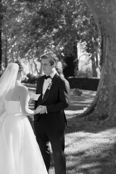 Private vows and vow reading as the couple face each other, hands clasped, sharing a handwritten vow card beneath garden trees by a large trunk.