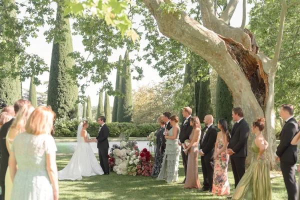 Ceremony moment at an outdoor wedding ceremony as the bride and groom hold hands beneath cypress trees, her long veil trailing on the lawn.