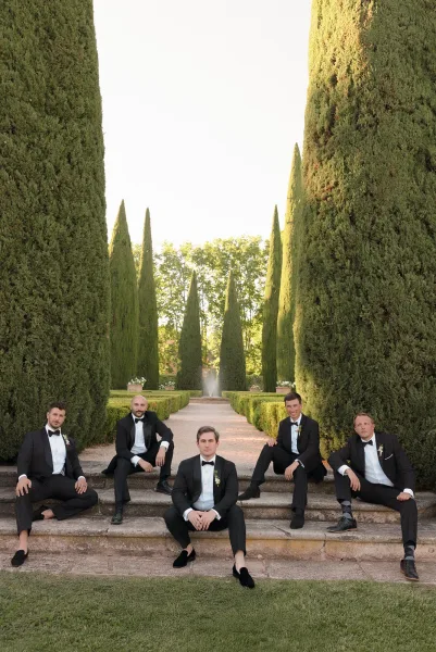 Groomsmen portrait of black tie groomsmen with bow ties, seated on stone steps in a formal garden framed by cypress trees and a fountain.