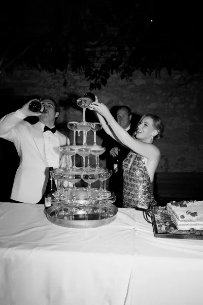 Champagne tower moment with the bride pouring champagne while the groom looks on beside a berry-topped wedding cake.
