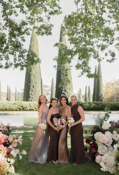 Bridesmaids portrait in mismatched bridesmaid dresses, holding bouquets by a reflecting pool amid cypress trees and hedges under an open sky.