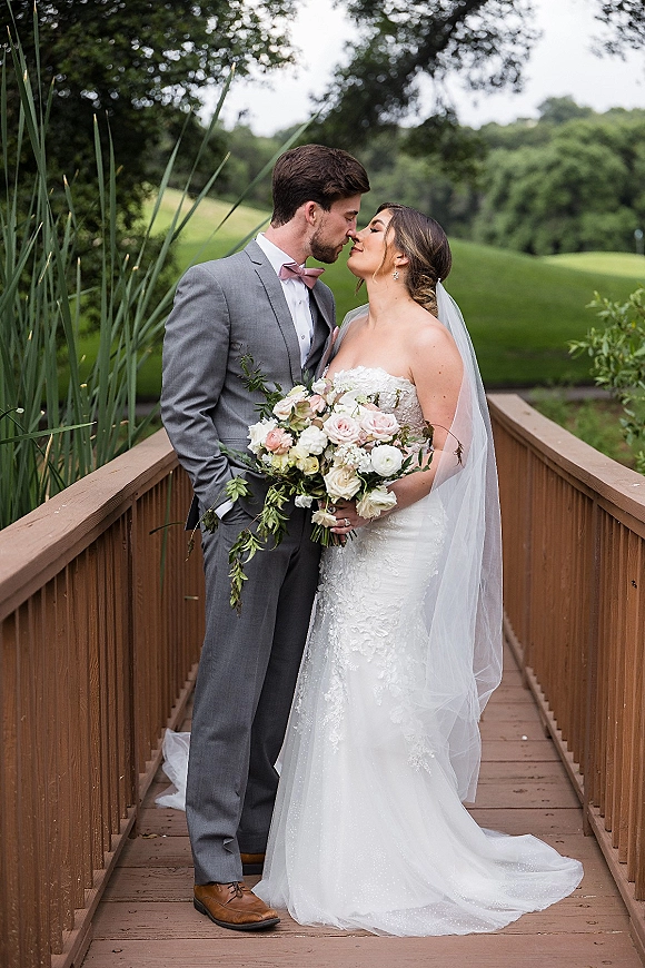 Wedding couple portrait of bride and groom kissing on a wooden bridge, bride holding a blush-and-white bouquet with veil over shoulder