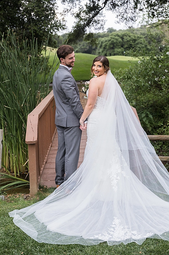 Couple portrait of bride and groom holding hands on a wooden footbridge, her long veil and lace gown trailing by reeds and hills