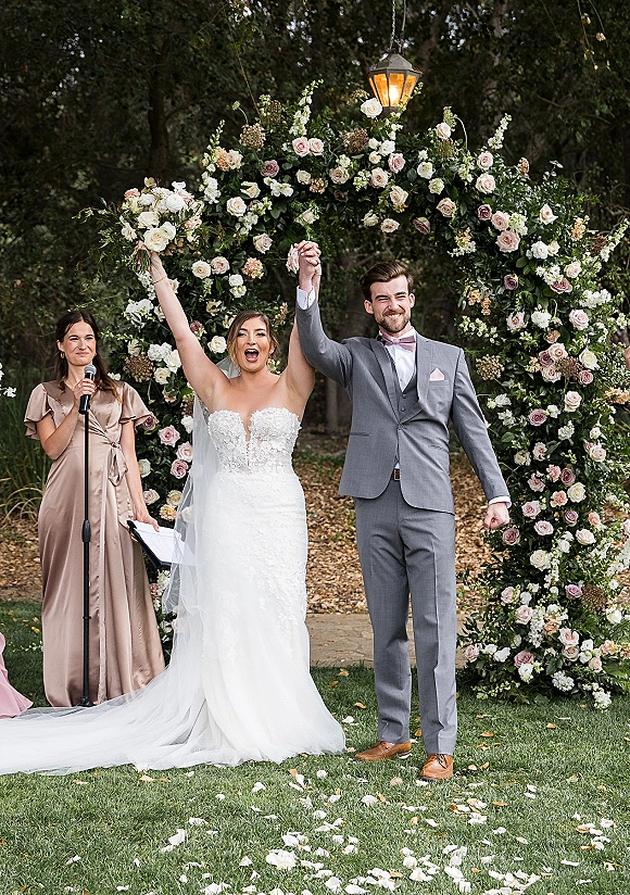 Recessional moment as bride and groom walking out with hands raised under a rose-and-greenery arch and hanging lantern on garden lawn