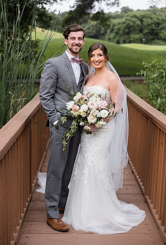 Couple portrait of bride and groom portrait on a wooden bridge, bride in strapless lace dress holding a blush rose bouquet amid greenery
