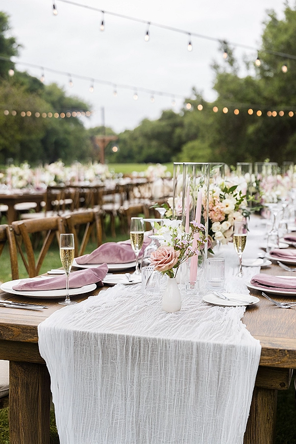 Reception tablescape with an outdoor wedding reception table on wood farm tables, blush napkins, floral centerpieces, and string lights overhead