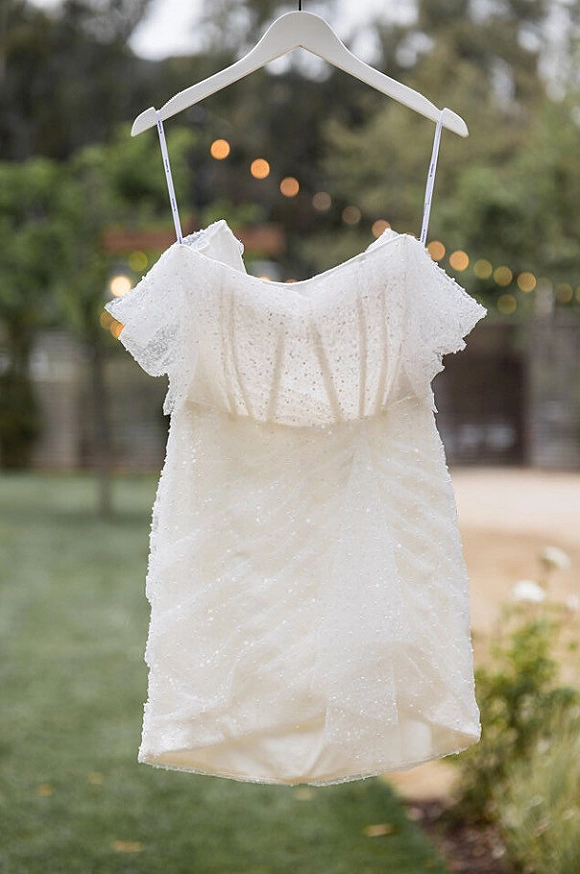 Wedding dress with beaded fabric on a hanger, spaghetti straps and off-shoulder sleeves, hanging outdoors by trees and string lights