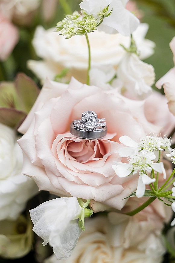 Wedding rings nestled in a blush rose bouquet, featuring a diamond halo engagement ring and dark metal band against soft white flowers