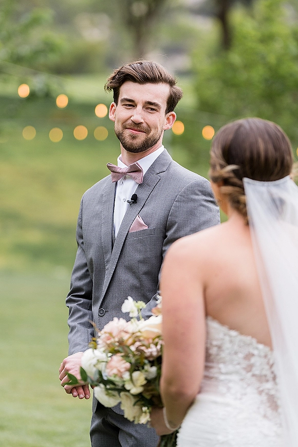 First look moment as bride approaches groom in a gray suit, her veil and bouquet visible on a garden lawn with string lights