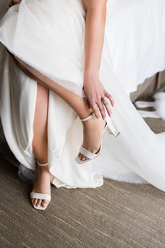 Bridal shoes and white wedding heels beside a bride’s manicured hand with engagement ring on a bed near the gown hem
