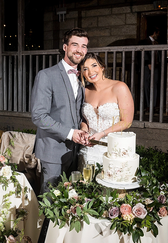 Cake cutting moment as bride in a strapless lace dress and groom in a gray suit slice a two tier cake with rose accents on a patio table