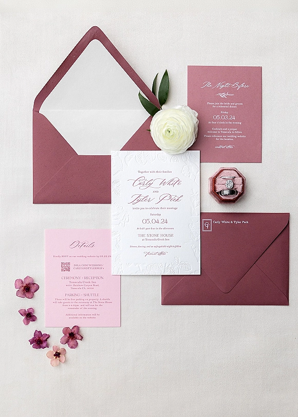 Invitation suite flatlay with burgundy envelopes, pink embossed cards, velvet ring box with rings, and ranunculus on a white backdrop