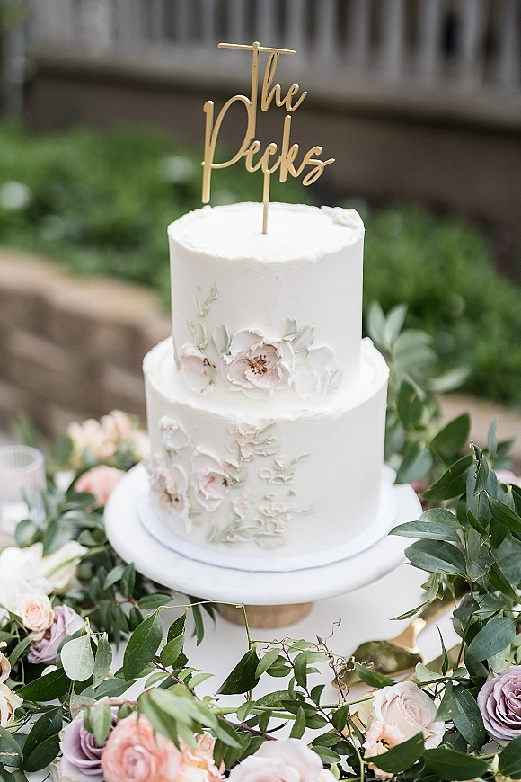 Wedding cake with white buttercream on a stand, topped with gold script and sugar flowers, set on a garden stone ledge with greenery
