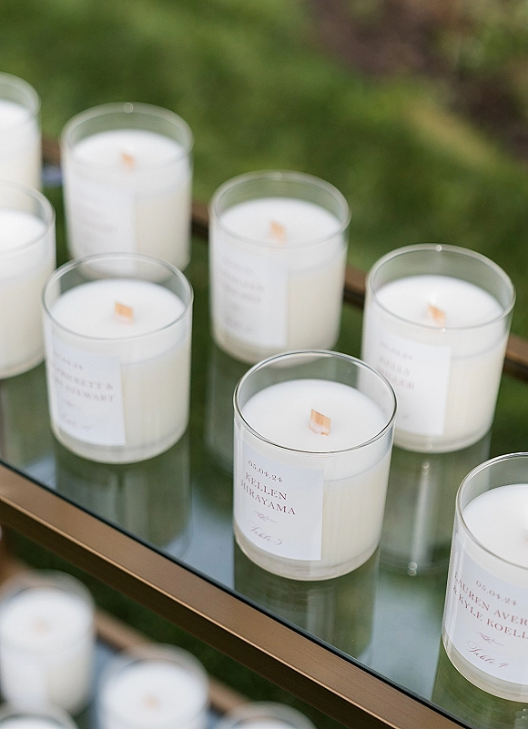 Wedding favor candles with personalized wedding favors labels in glass jars on a mirrored tray with a gold frame, set on a green lawn