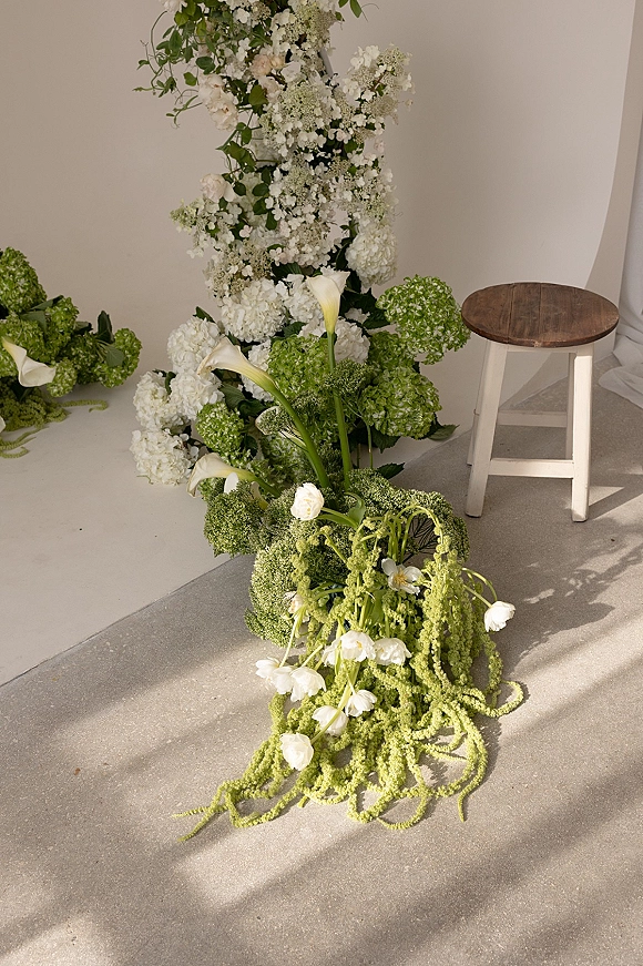 Wedding ceremony flowers in a ceremony floral installation of hydrangeas and calla lilies with greenery cascading on a floral foam base, against a white wall