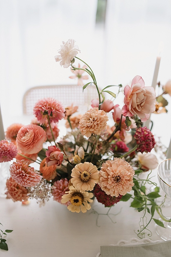 Wedding centerpiece with a floral table centerpiece of blush and coral blooms, greenery, taper candles, and clear goblet on a white tablecloth by bright windows